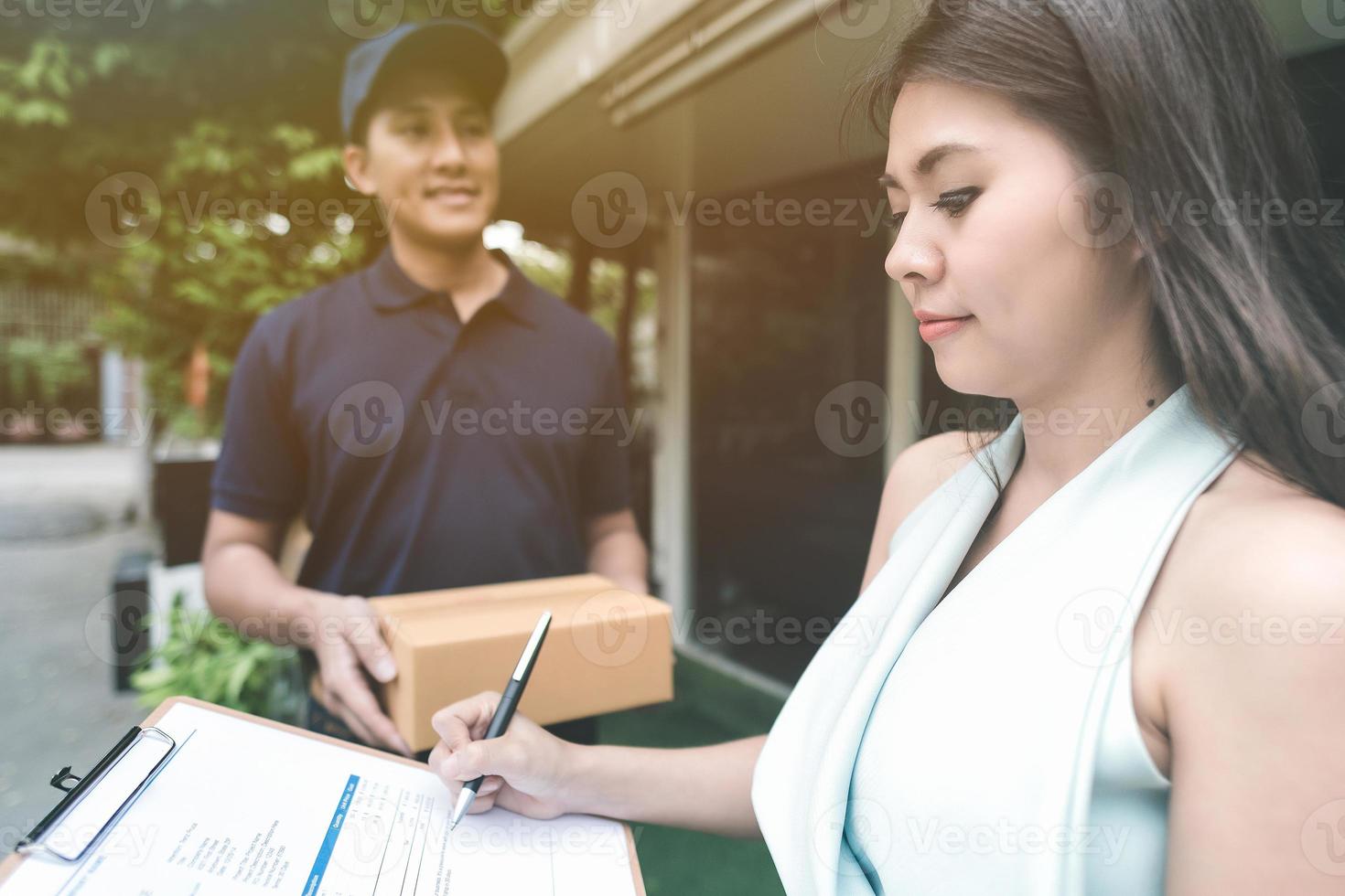 knappe jonge aziatische bezorger die lacht en een kartonnen doos vasthoudt terwijl hij wacht mooie jonge aziatische vrouw die handtekening zet in slimme tablet voor bevestiging van ontvangst. foto