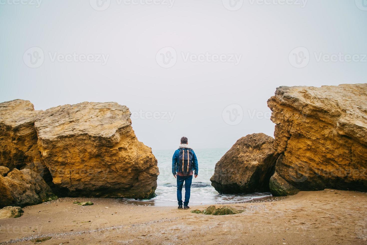 reiziger man met rugzak staande op zandstrand in het midden van rotsen tegen de achtergrond van de zee foto