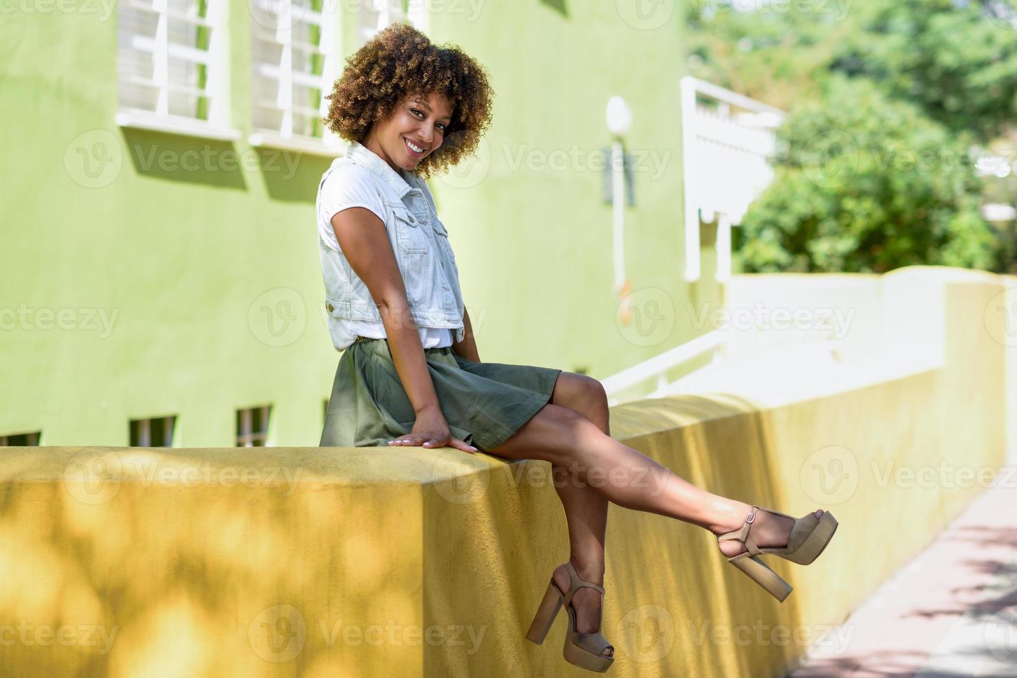 jonge zwarte vrouw, afrokapsel, zittend op een stadsmuur foto