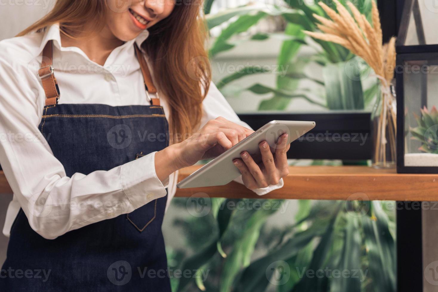 jonge aziatische vrouwelijke barista die elektronische tablet gebruikt in haar eigen coffeeshop terwijl ze een pauze neemt in de warme middag. jonge vrouwelijke barista en haar moderne kleine koffieshop. concept van de voedsel- en drankenindustrie. foto