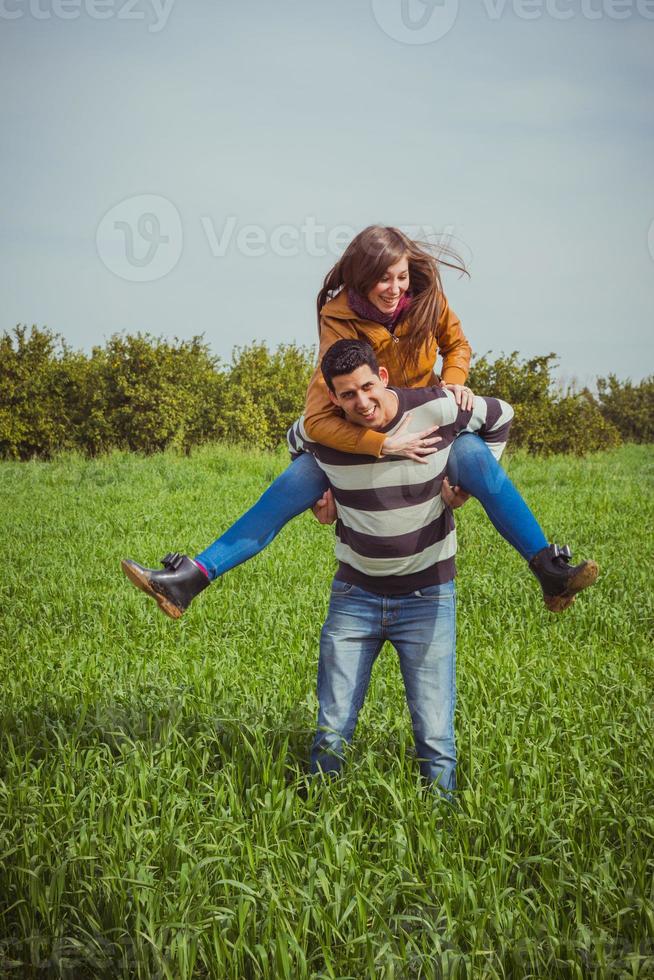 jong koppel plezier samen in groene veld. de vrouw die op de schouders van de man rijdt foto