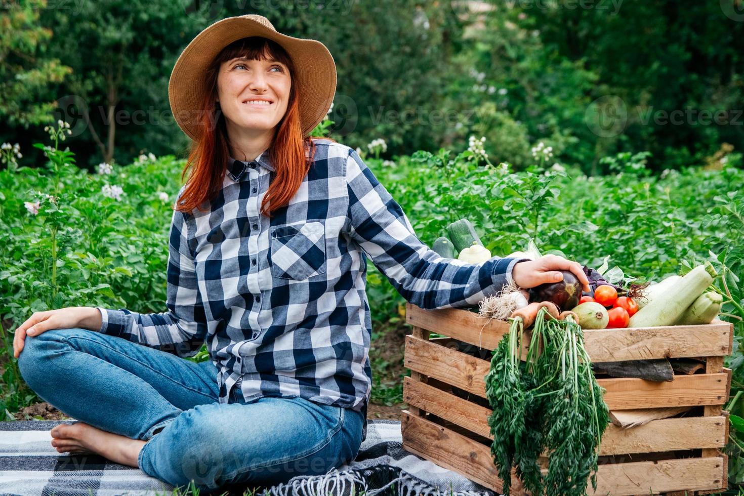 vrouw boer zittend op een deken in de buurt van verse biologische groenten in een houten kist tegen de achtergrond van een moestuin foto