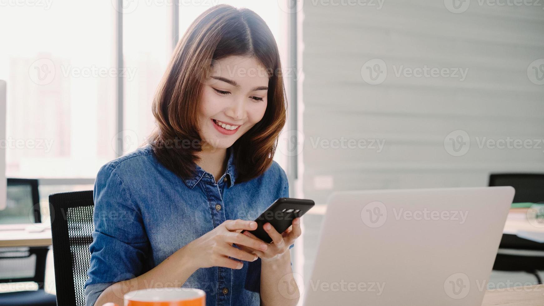 mooie jonge glimlachende aziatische vrouw die op laptop werkt terwijl ze geniet van het gebruik van smartphone op kantoor. Aziatische zakenvrouw werkdocument financiën in kantoor aan huis. genieten van tijd op kantoor. foto