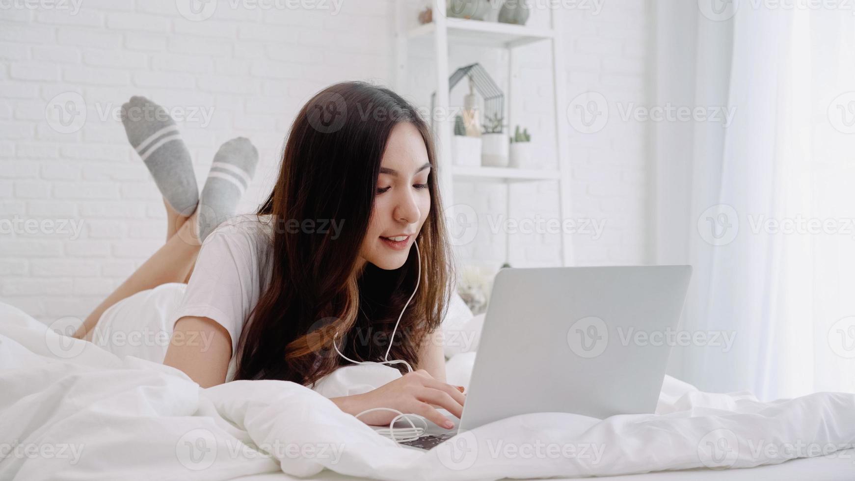 portret van mooie aantrekkelijke aziatische vrouw die computer of laptop gebruikt en muziek luistert terwijl ze op het bed ligt wanneer ze thuis in haar slaapkamer ontspant. levensstijl vrouwen die ontspannen tijd thuis concept gebruiken. foto