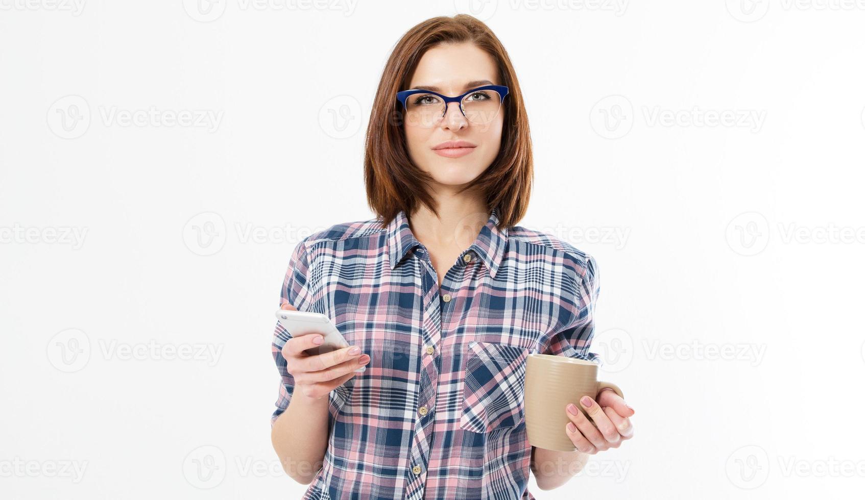 mooie jonge vrouw die een smartphone vasthoudt, een apparaat gebruikt, een stijlvolle bril draagt, glimlacht, een mok vasthoudt, geïsoleerd op een witte achtergrond, spijkerhemd foto