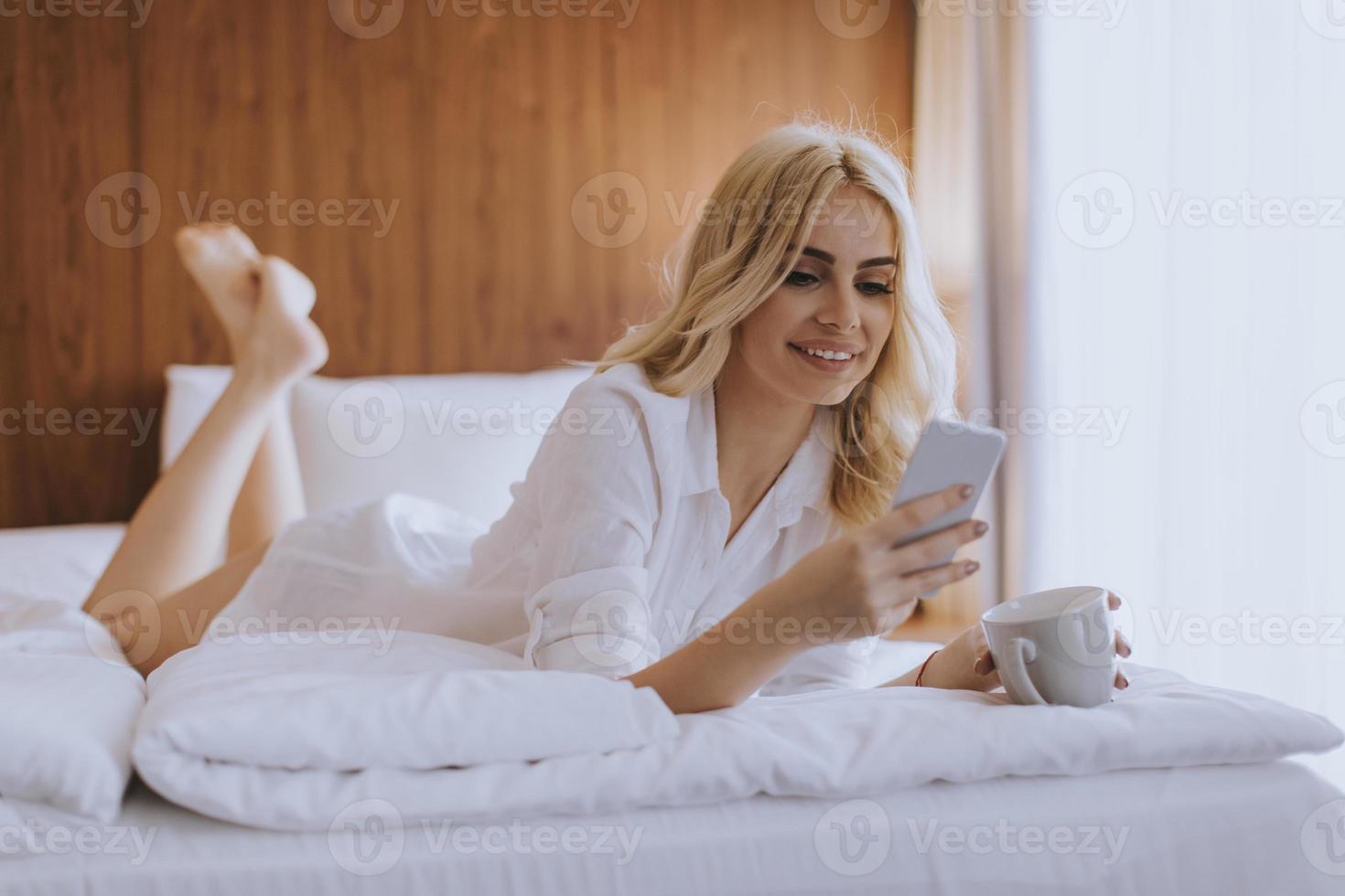 gelukkige vrouw die een mobiele telefoon gebruikt terwijl ze op bed ligt en 's ochtends koffie drinkt foto