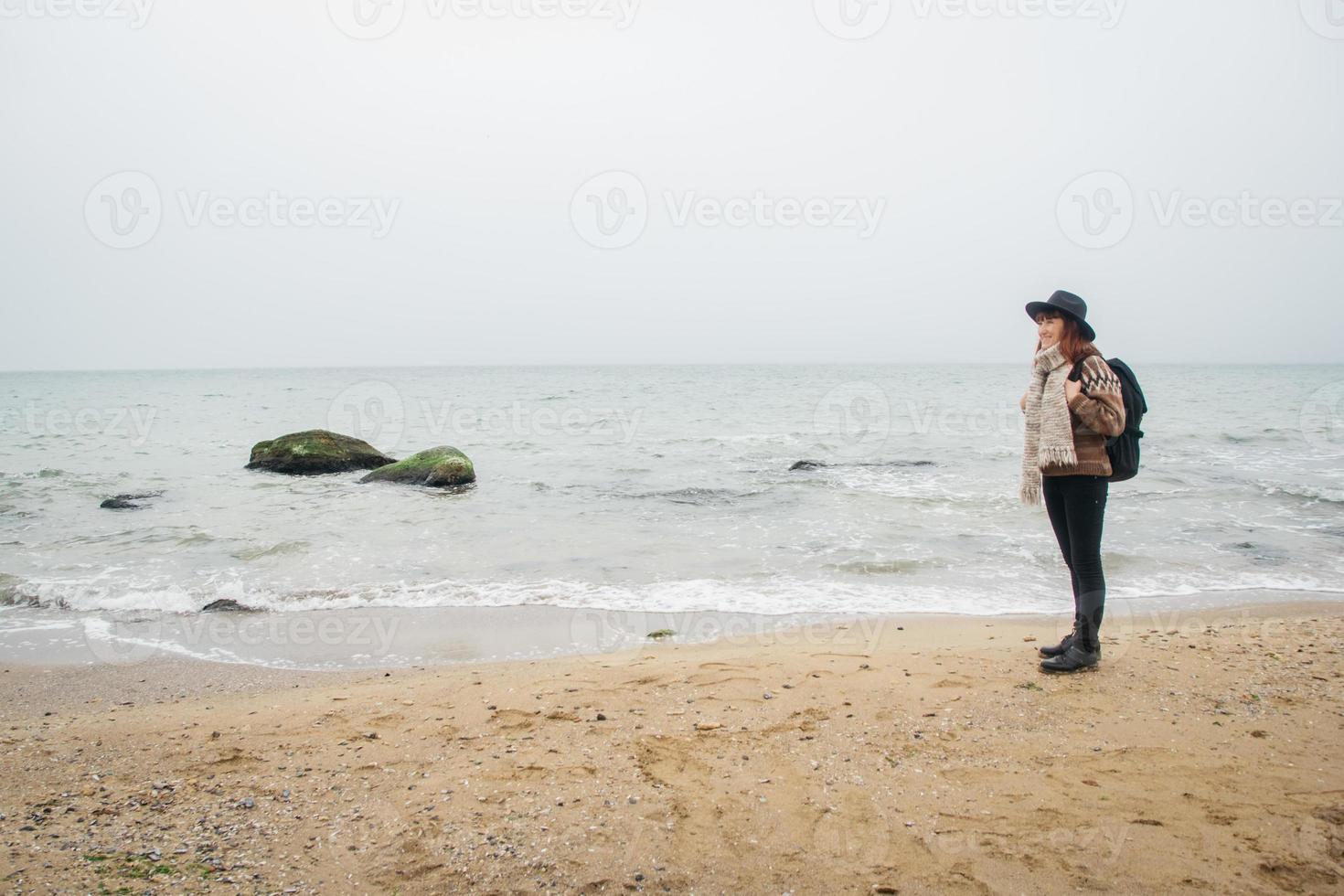 mooie roodharige vrouw in een muts en sjaal met een rugzak aan de kust tegen de achtergrond van de rotsen tegen de prachtige zee foto