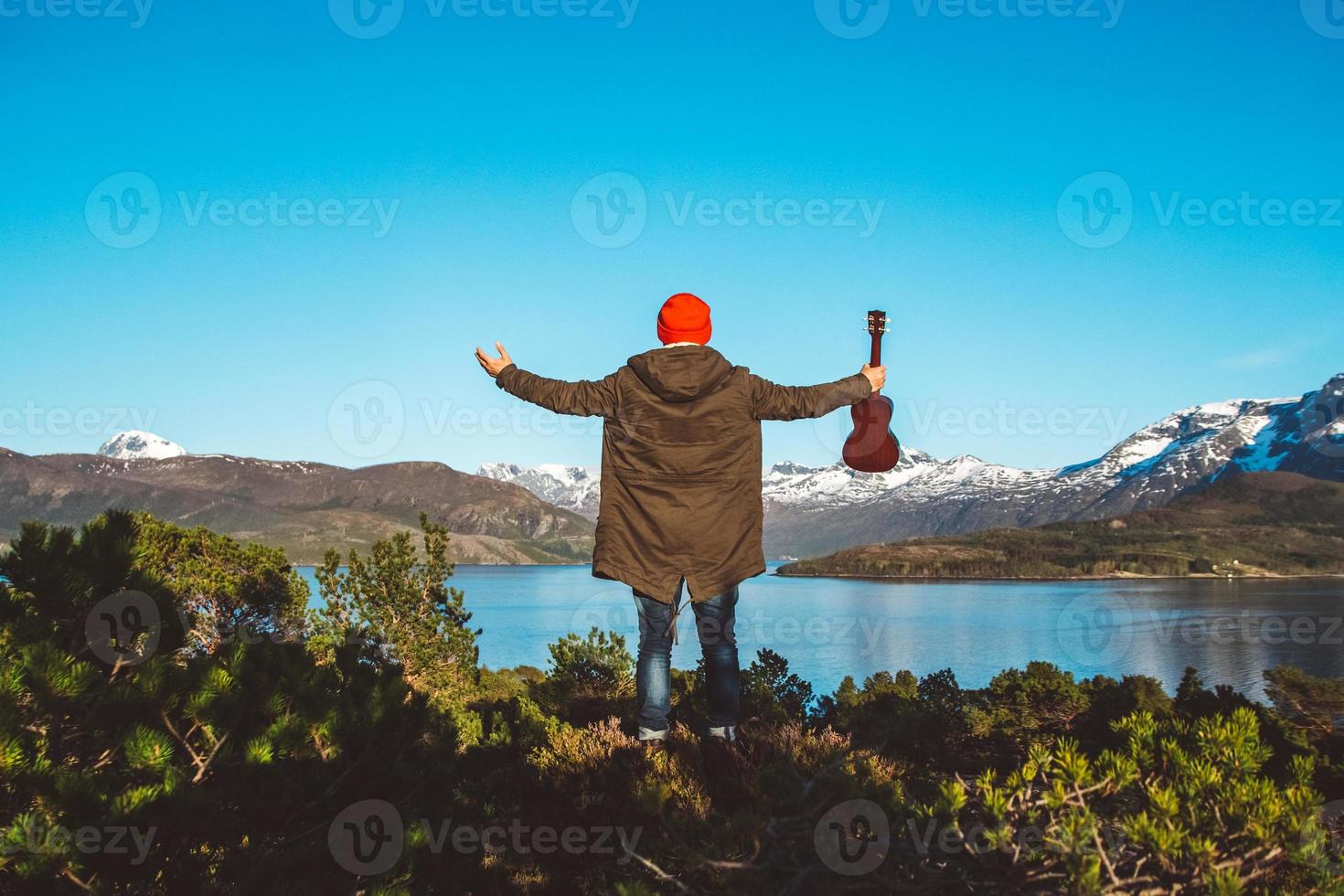 reiziger man staat in het midden van een bos met een gitaar op de achtergrond van bergen en meer. plaats voor tekst of reclame. van achteren schieten foto