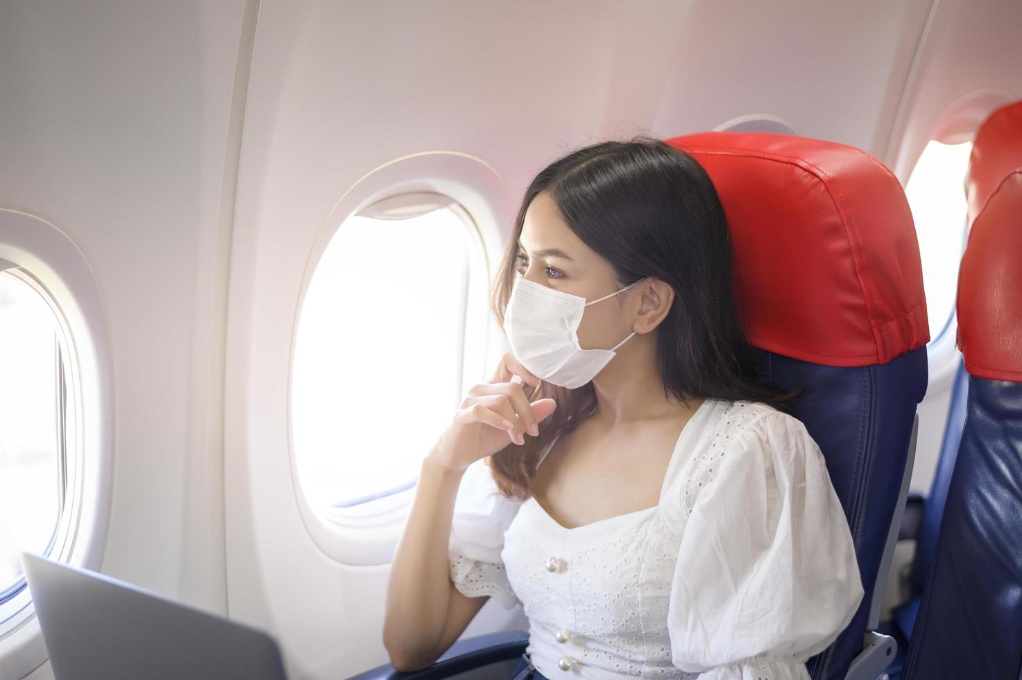 een jonge vrouw met een gezichtsmasker gebruikt laptop aan boord, nieuw normaal reizen na covid-19 pandemisch concept foto