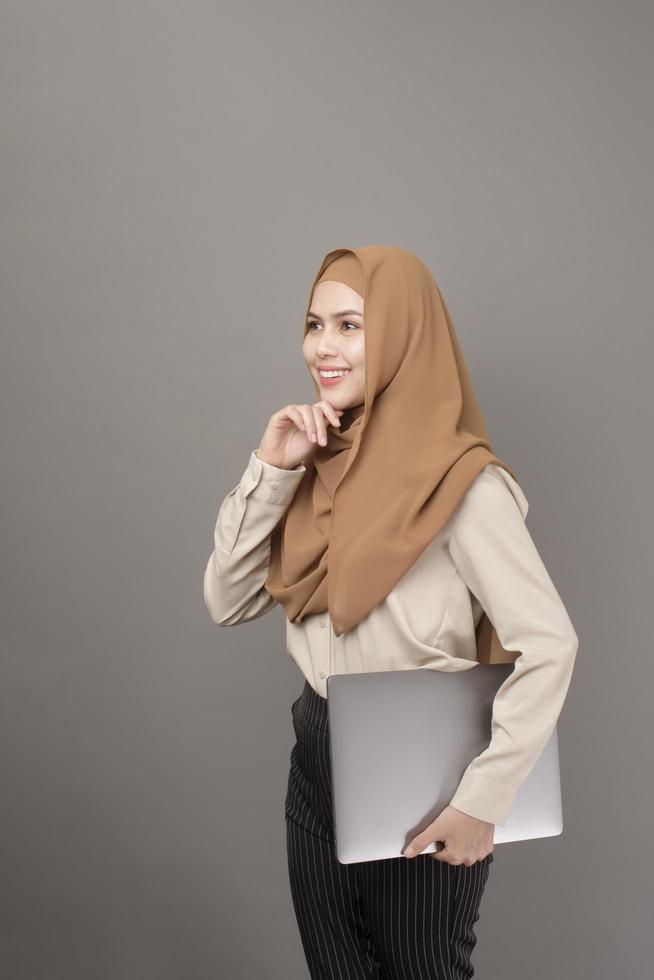 portret van mooie vrouw met hijab houdt computerlaptop op grijze achtergrond foto
