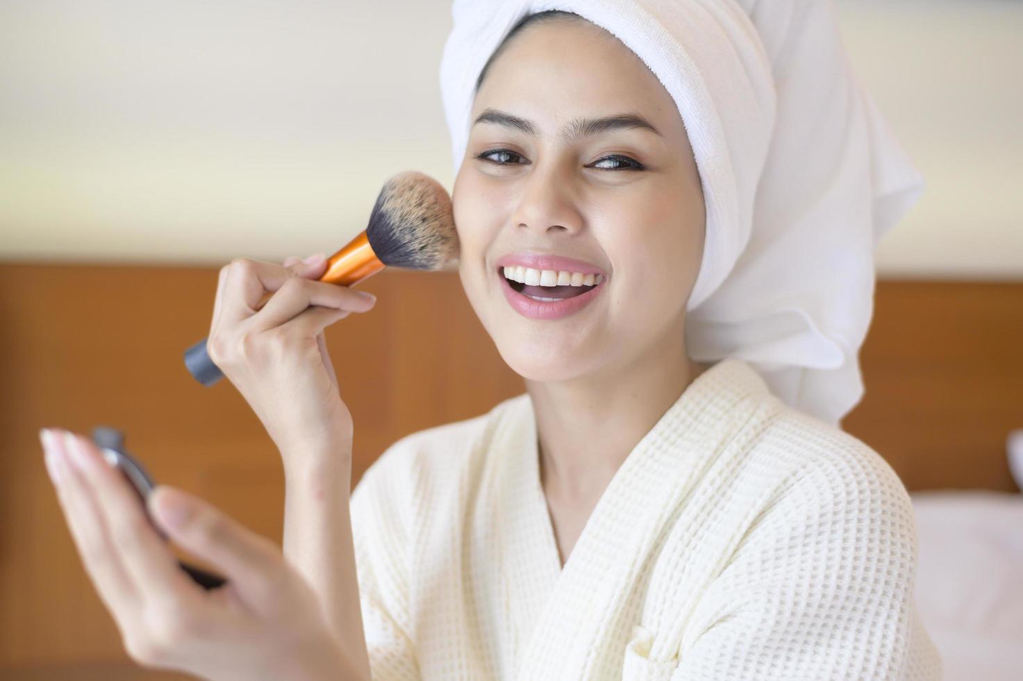 aantrekkelijke gelukkige vrouw in witte badjas past natuurlijke make-up toe met cosmetische poederborstel, schoonheidsconcept. foto
