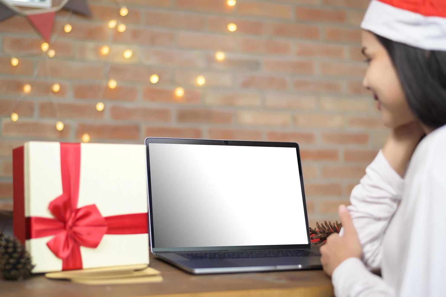 jonge lachende vrouw met rode kerstman hoed video bellen op sociaal netwerk met familie en vrienden op eerste kerstdag. laptop monitorscherm mock-up. foto