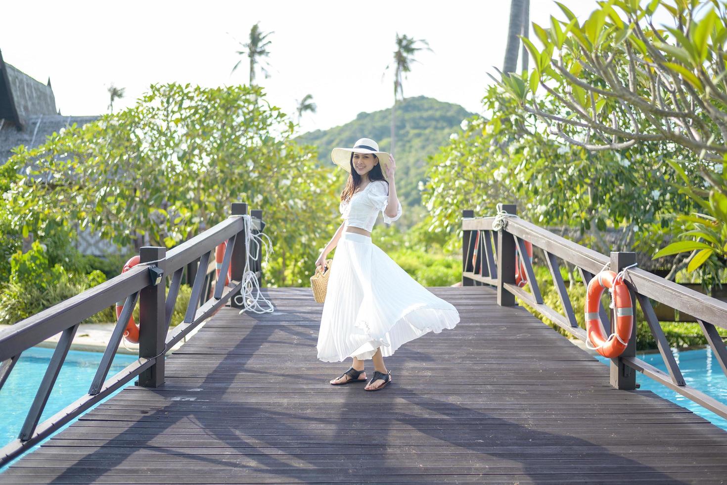 een gelukkige mooie vrouw in witte jurk die geniet van en staat op een houten brug boven het zwembad in een gezellige bungalow met groene tropische tuin op het eiland phi phi, thailand foto