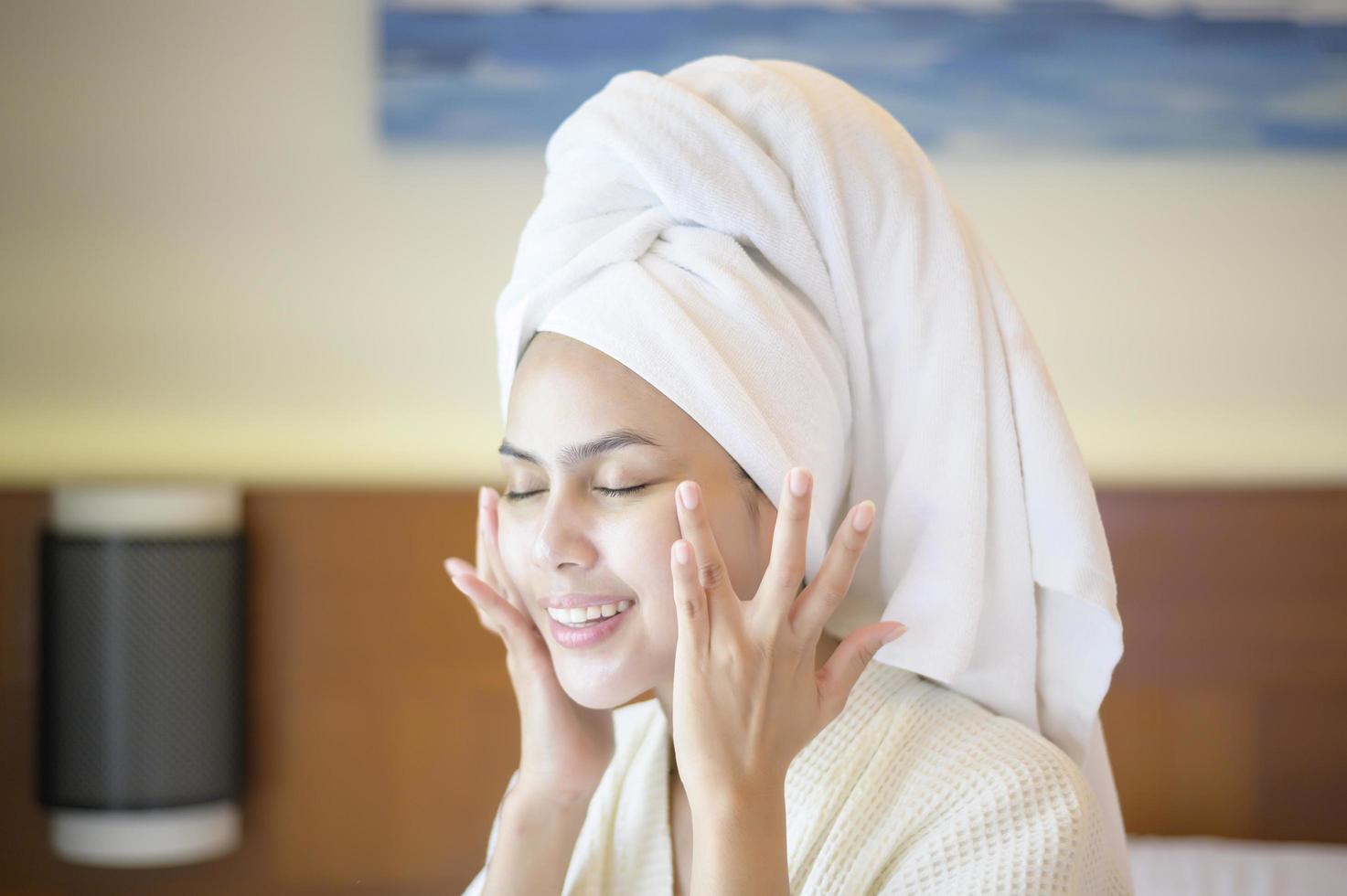 een gelukkige mooie vrouw in witte badjas die vochtinbrengende crème op het gezicht aanbrengt in de slaapkamer, huidverzorging en behandelingsconcept foto
