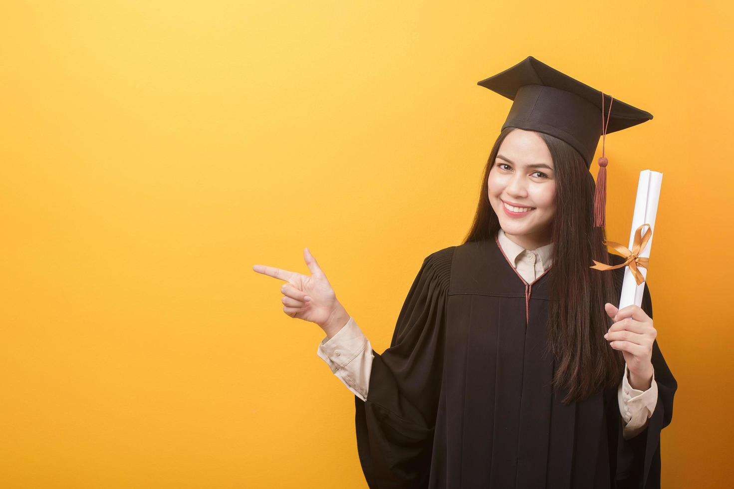 portret van gelukkige mooie vrouw in afstudeerjurk houdt onderwijscertificaat op gele achtergrond foto