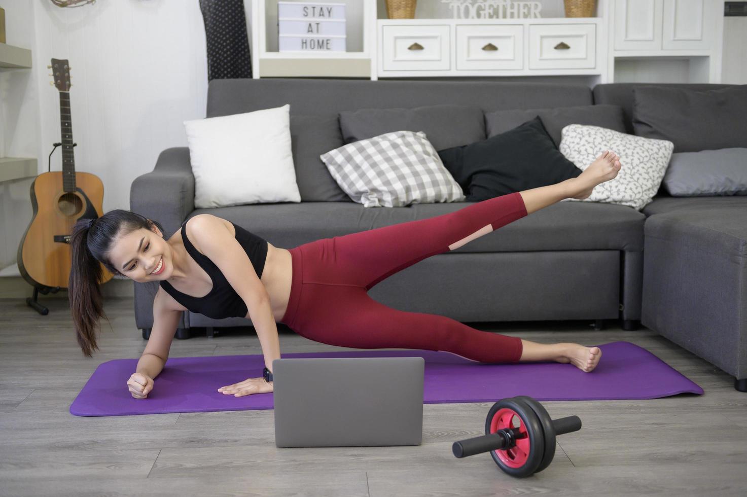 een vrouw doet yogaplank en kijkt naar online trainingshandleidingen op haar laptop in de woonkamer, fitnesstraining thuis, concept voor gezondheidszorgtechnologie. foto