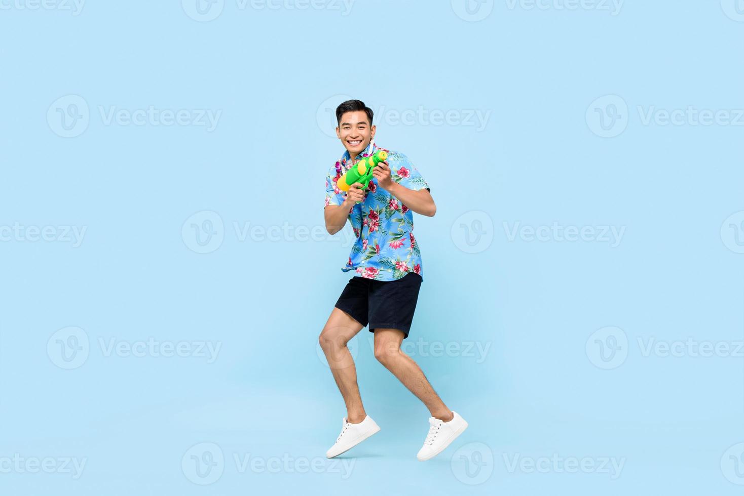 knappe glimlachende jonge aziatische man spelen met waterpistool en springen in studio blauwe achtergrond voor songkran festival in thailand en zuidoost-azië foto