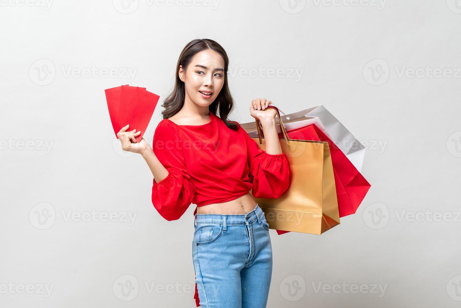 Glimlachende gelukkige Aziatische vrouw met boodschappentassen en rode enveloppen geïsoleerd in lichtgrijze studio achtergrond voor Chinees Nieuwjaar verkoop concept foto