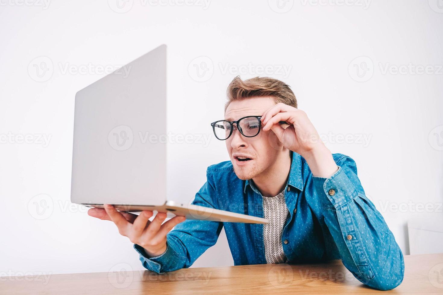 man in shirt en bril kijkt hij naar laptop met verbazing en met een grappig gezicht aan tafel op een witte achtergrond foto
