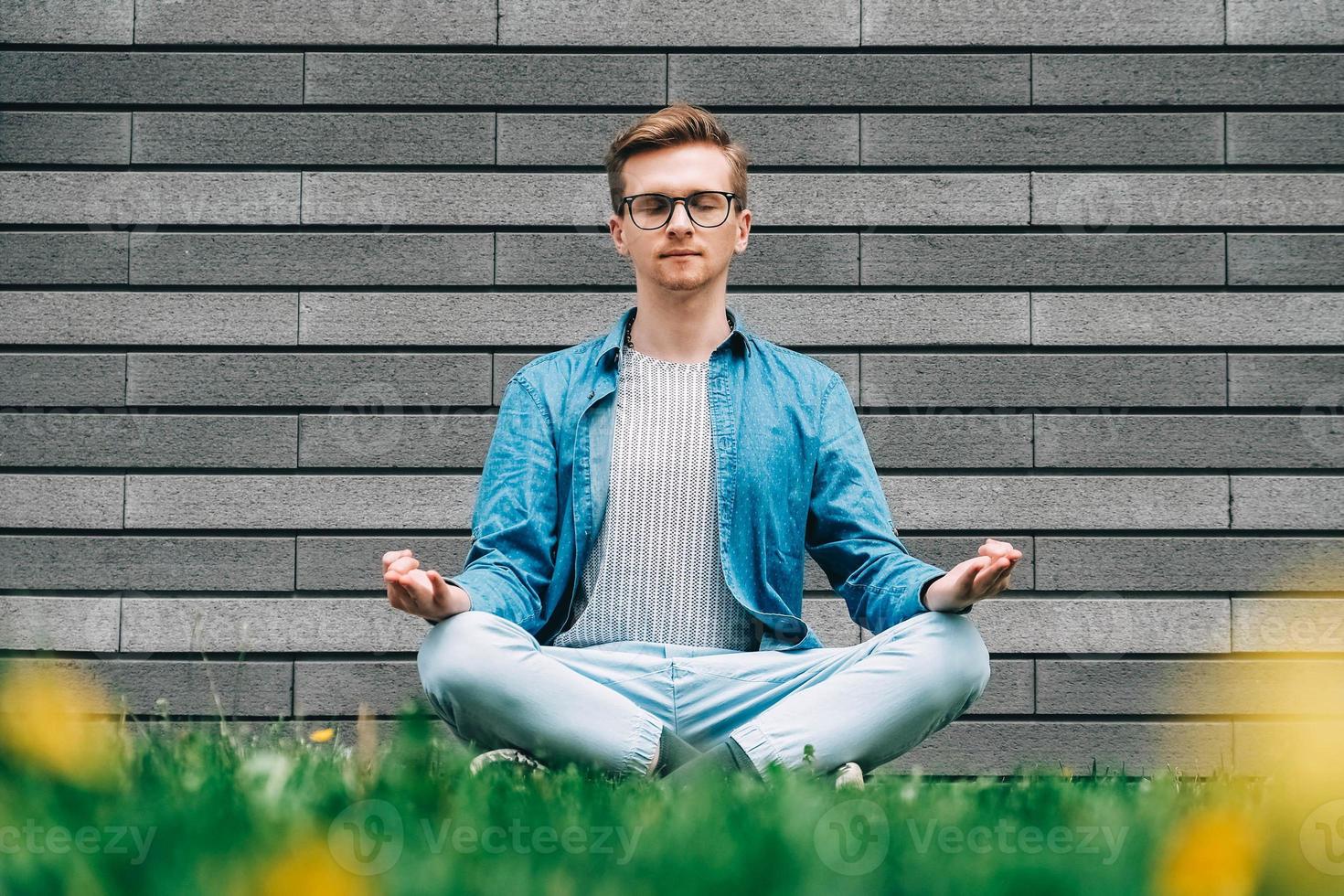 man in shirt, spijkerbroek en bril zittend in pose van meditatie op groen gras op een achtergrond van een grijze muur foto