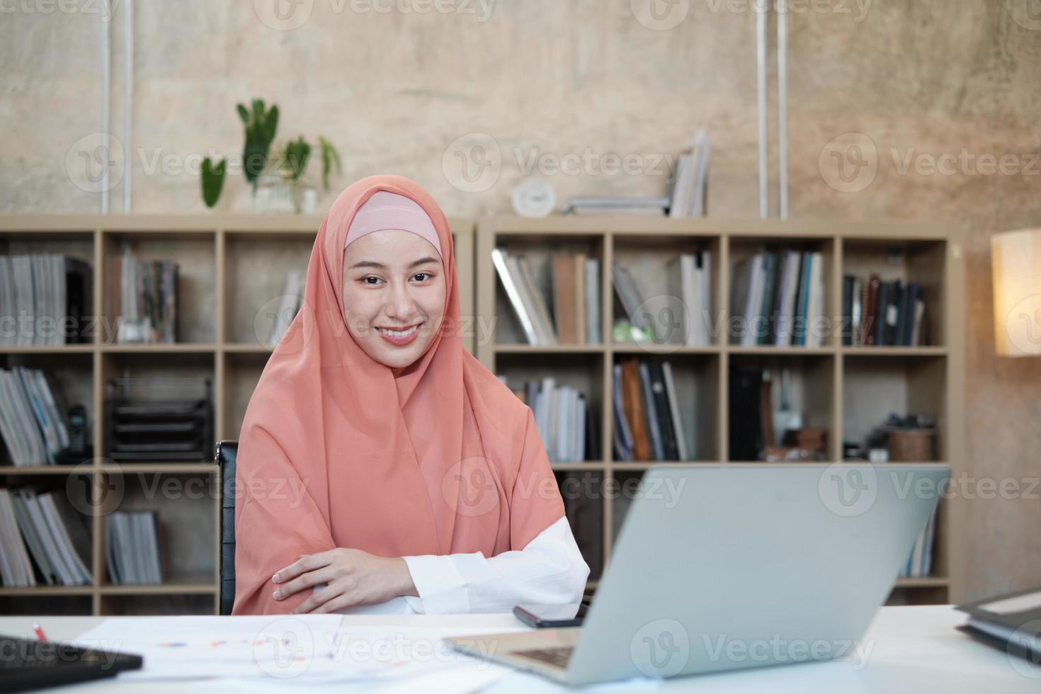 portret van de mooie oprichter van het startende bedrijf die een islamitische vrouwelijke persoon met hijab is, glimlacht en kijkt naar de camera in een kleine kantoorwerkplek, werkend met een laptop op een wit bureau, een boekenplank achter. foto