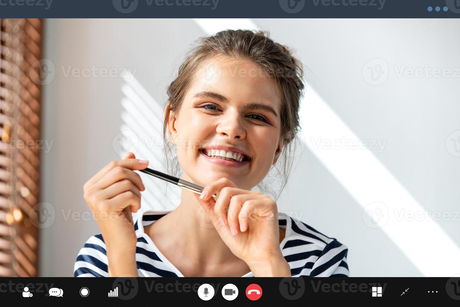 Glimlachende blanke vrouw die een pen vasthoudt en naar de camera kijkt terwijl ze een videoconferentiegesprek voert in de woonkamer, leert en werkt vanuit huisconcept foto