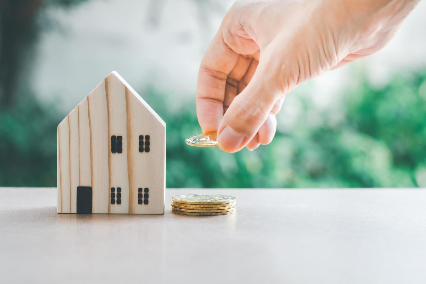 geld besparen concept. hand houden gloden munt met houten modelwoning op tafel met groene bokeh achtergrond. sparen of beleggen voor thuis in de toekomst. foto