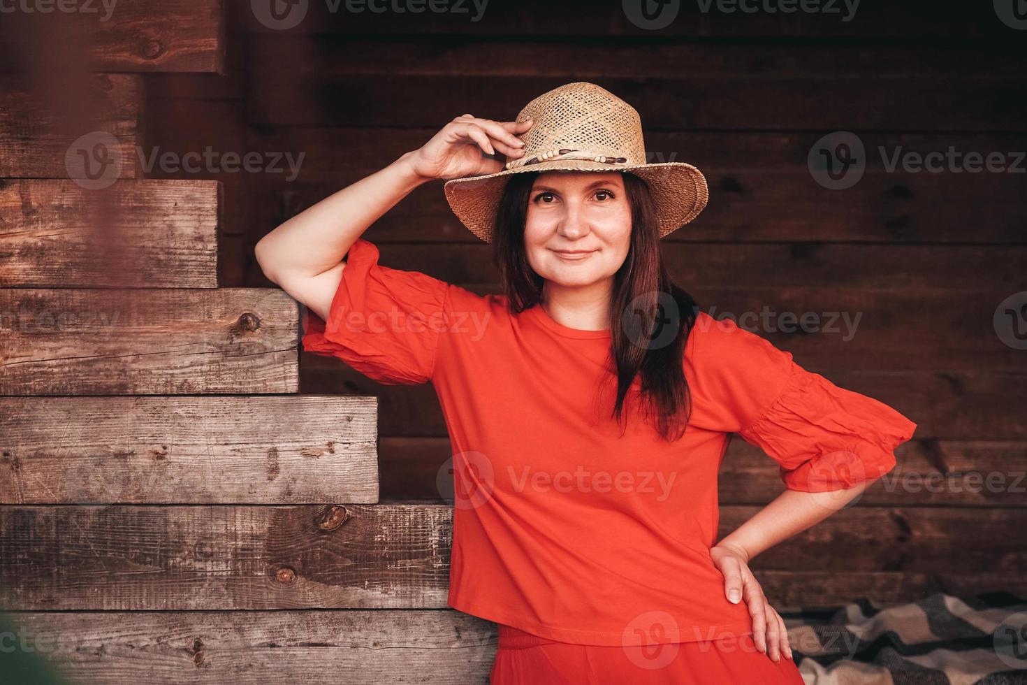 portret van een mooie vrouw met een strohoed gekleed in rode kleren staat op de achtergrond van een houten huis foto