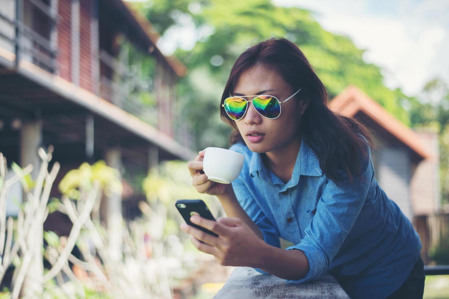 jonge hipster vrouw kijkt naar haar telefoon terwijl ze 's ochtends een kopje koffie vasthoudt en glimlacht. ontspannen vakantieconcept. foto