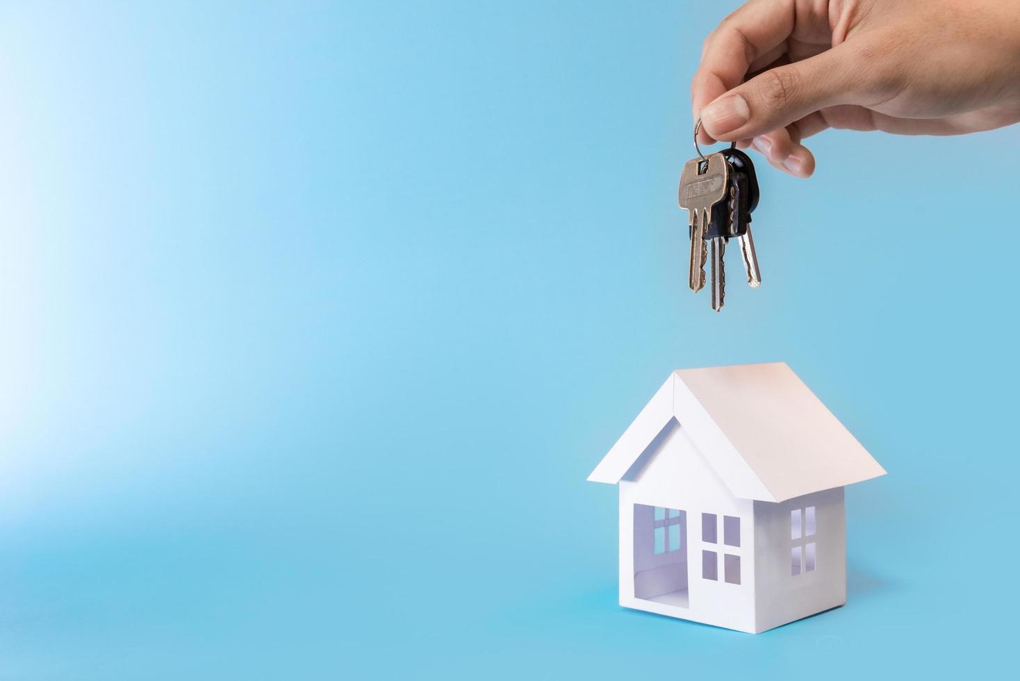 hand met sleutels en witte huis model op blauwe achtergrond voor herfinancieren plan en onroerend goed concept. foto