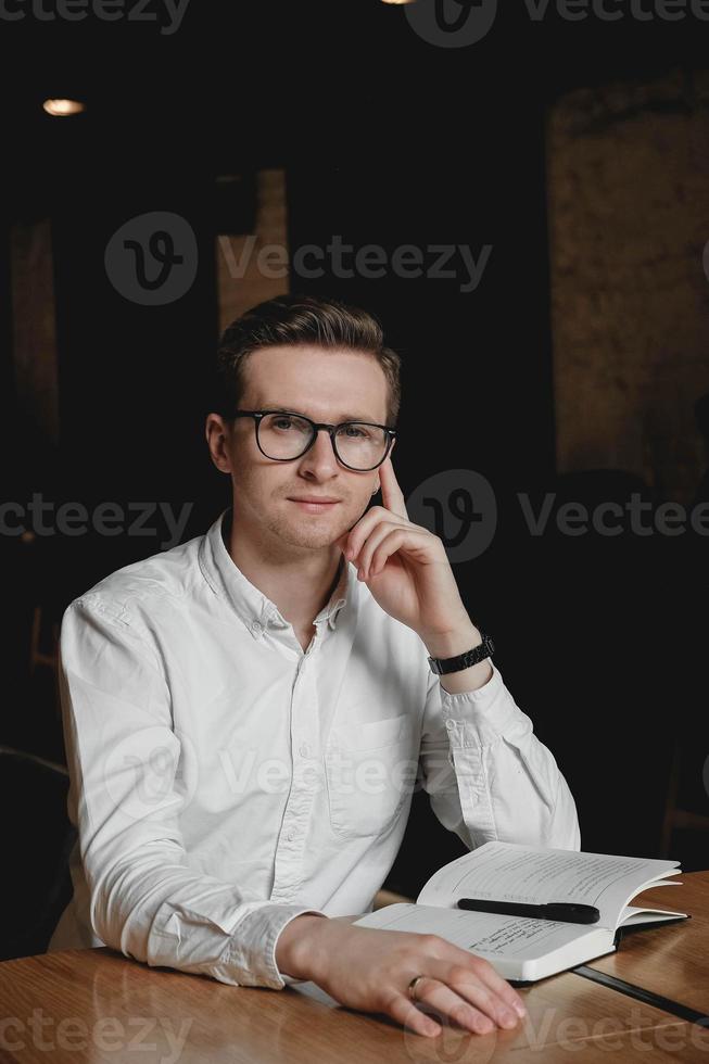 jonge man met een wit overhemd en een bril die aan de tafel zit en een pen vasthoudt, maakt aantekeningen op zijn agenda op een donkere achtergrond op kantoor. kopiëren, lege ruimte voor tekst foto