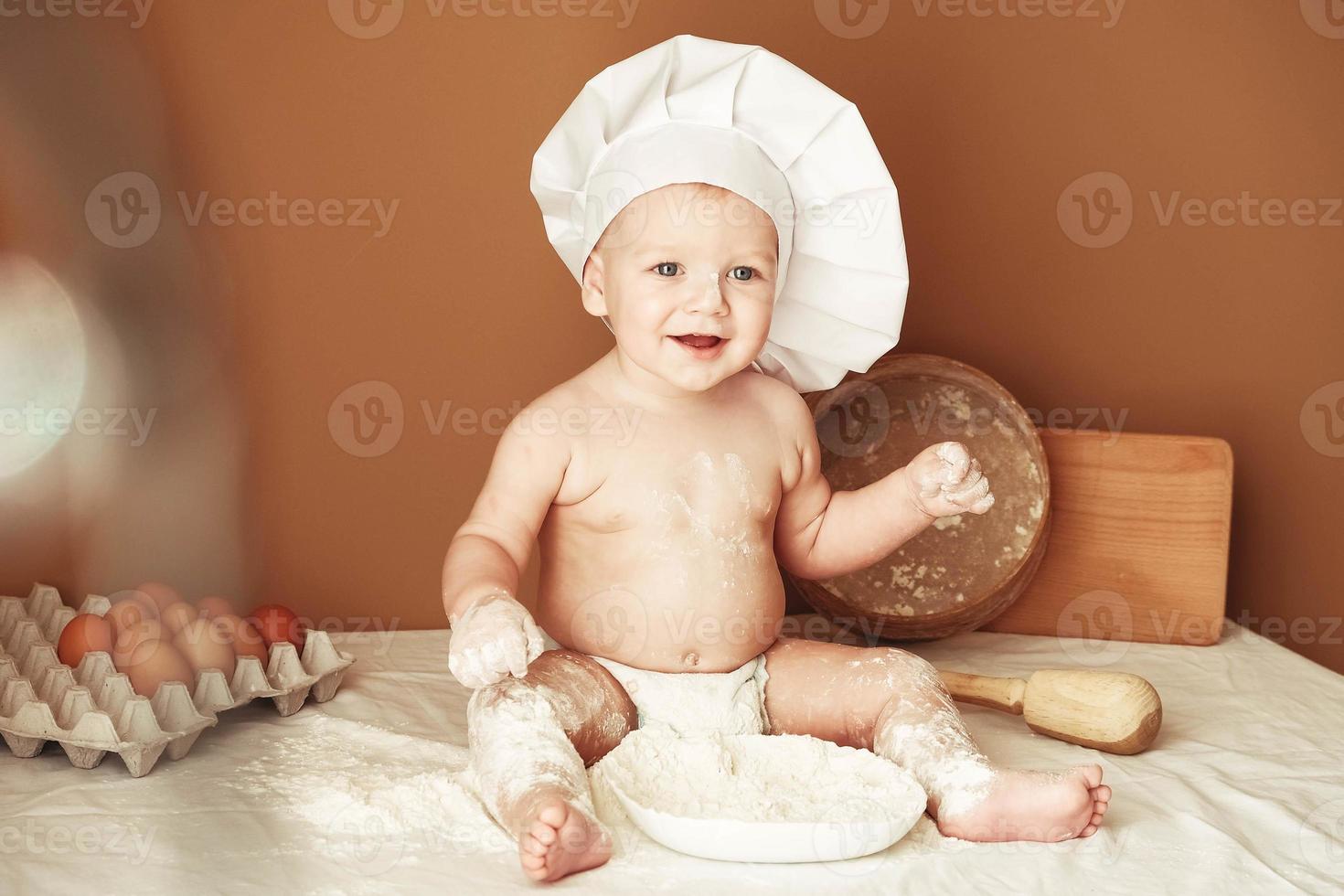 kleine jongen bakker in een koksmuts zittend op de tafel spelen met bloem op een bruine achtergrond met een houten deegroller, een ronde rustieke zeef en eieren. kopiëren, lege ruimte voor tekst foto