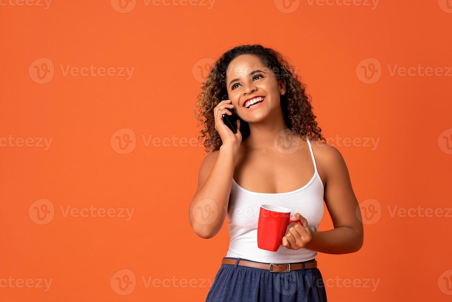 Gelukkig lachende Afro-Amerikaanse vrouw bellen op mobiele telefoon met koffiekopje in de hand geïsoleerd op oranje studio achtergrond foto