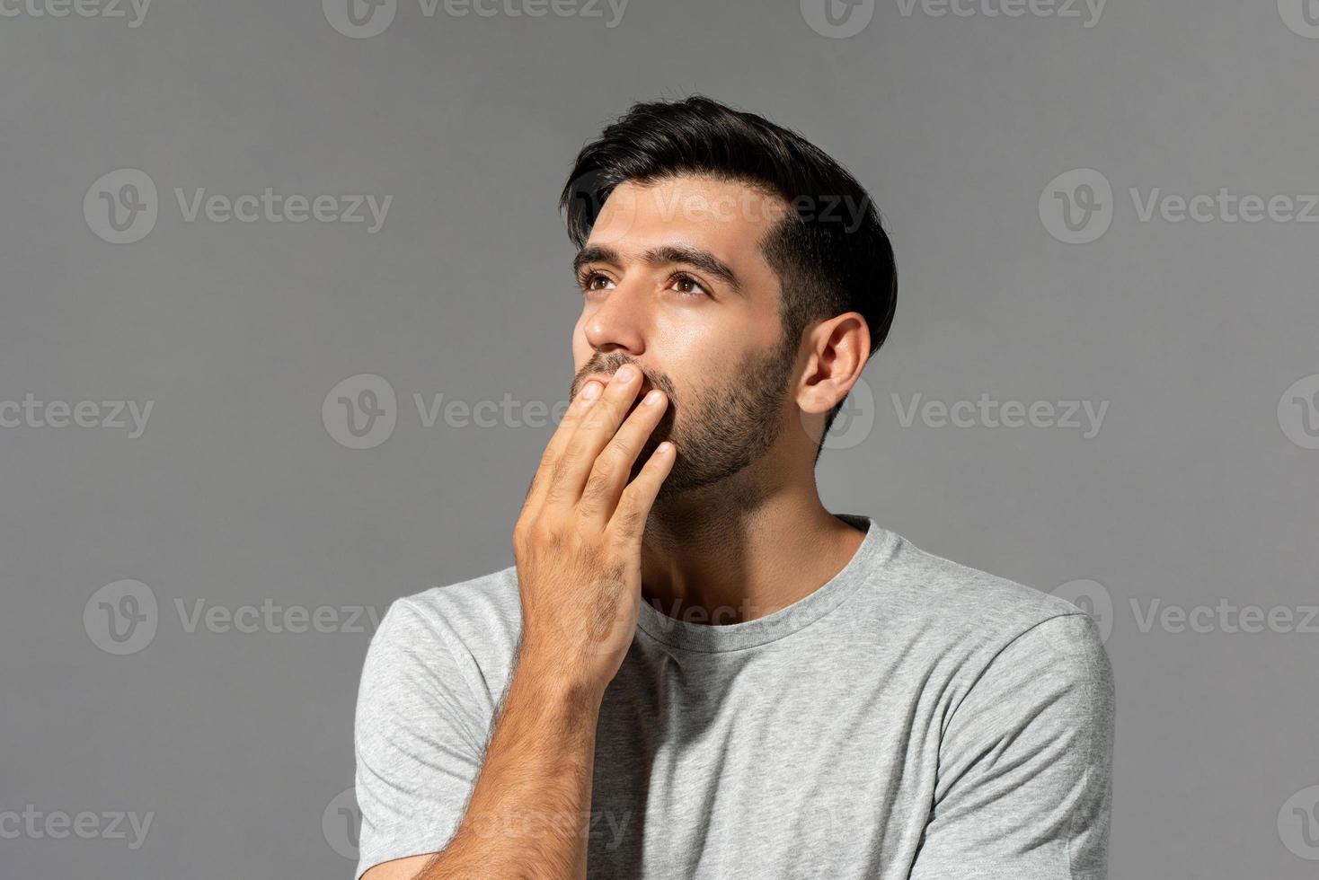 geschokte jonge blanke man die opkijkt en denkt met de mond van de handbedekking op een lichtgrijze studioachtergrond foto