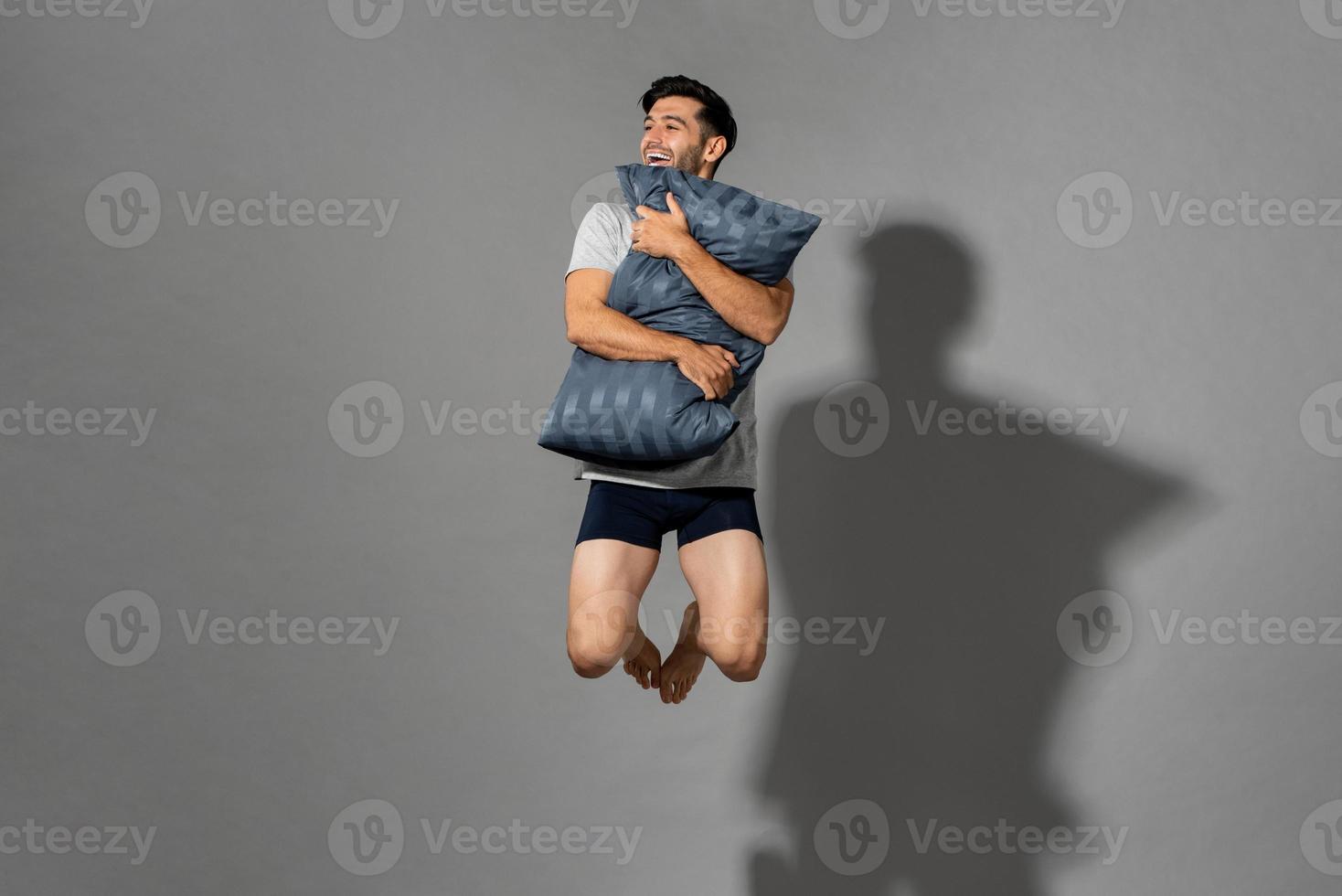 portret van een jonge, frisse, energieke man met nachtkleding die een kussen vasthoudt en in de lucht springt nadat hij 's ochtends wakker is geworden uit een goede nachtrust, studio-opname op een grijze geïsoleerde achtergrond foto