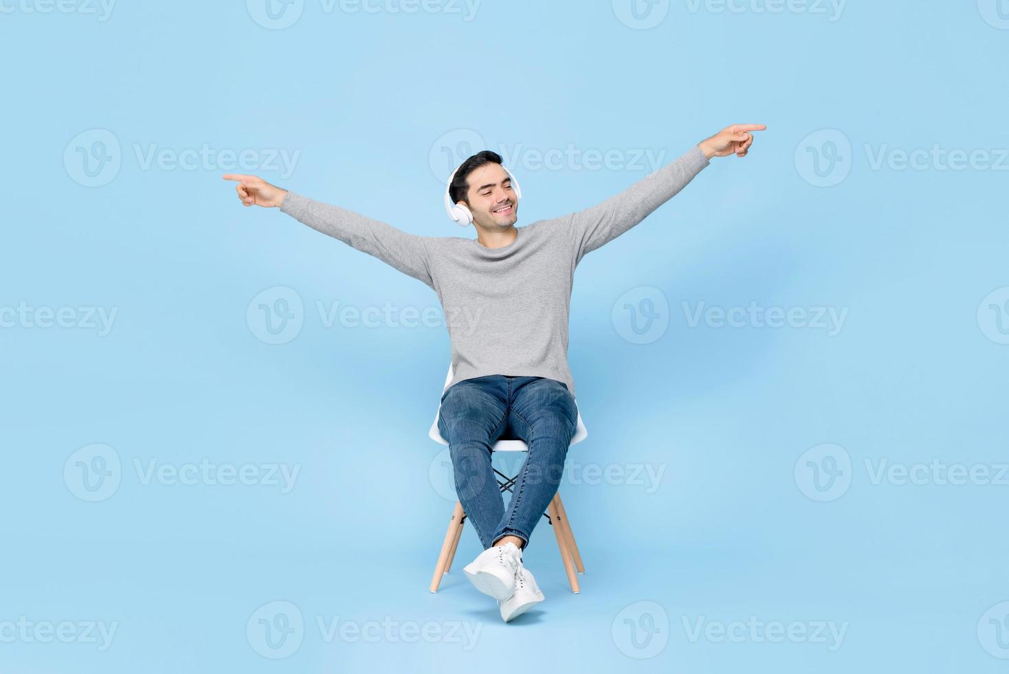 portret van een ontspannende jonge knappe blanke man zittend op een stoel met een koptelefoon op, luisterend naar muziek met opgeheven armen in een geïsoleerde studio blauwe achtergrond foto