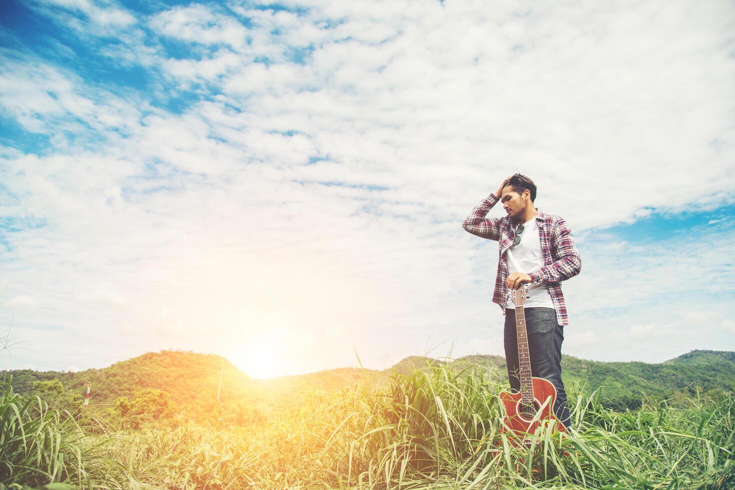 jonge hipster man met een gitaar met een wandeling in de natuur, ontspannen in het veld in een zonnige blauwe hemeldag. foto