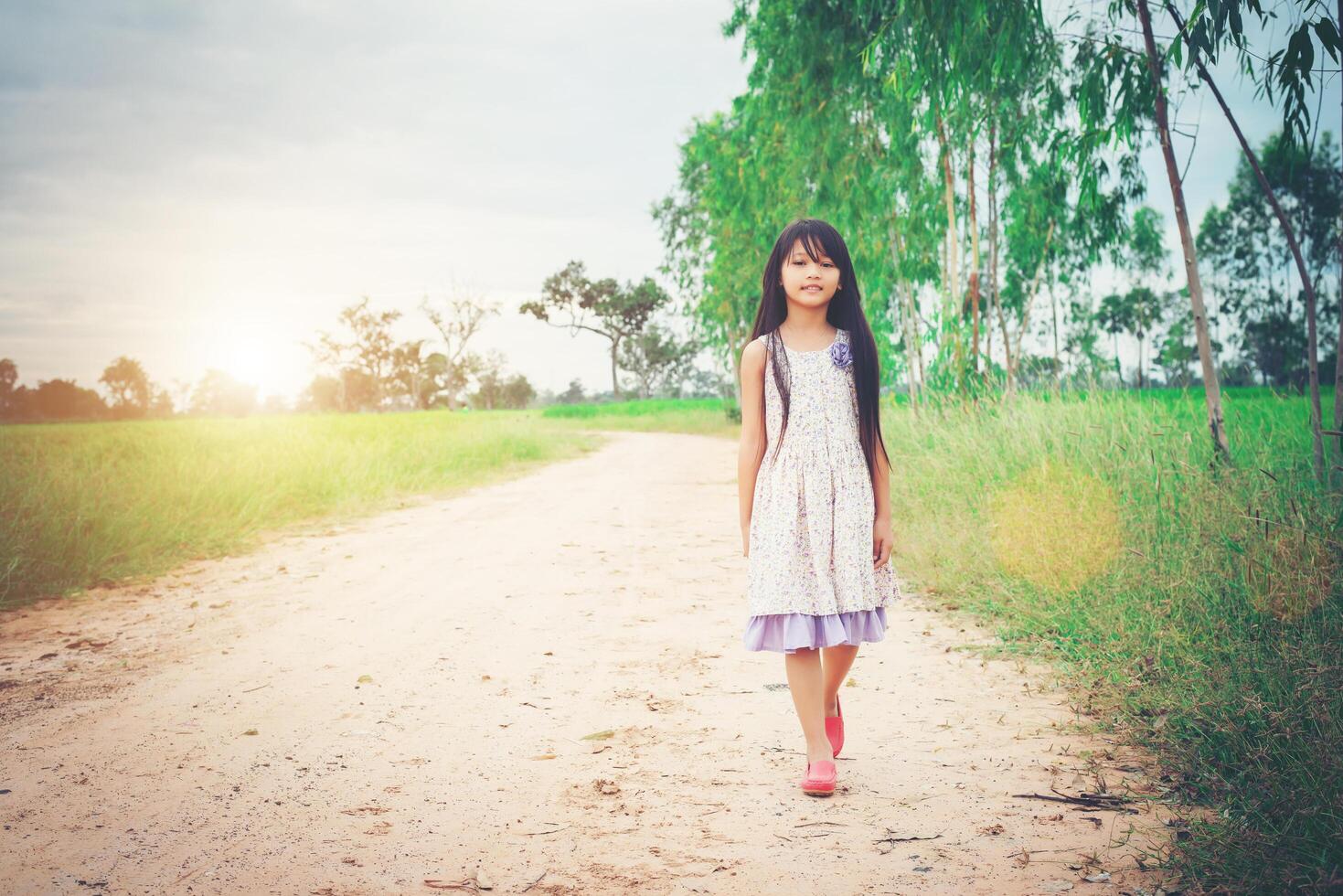 klein meisje met lang haar dat een jurk draagt, loopt van je weg op een landelijke weg. foto