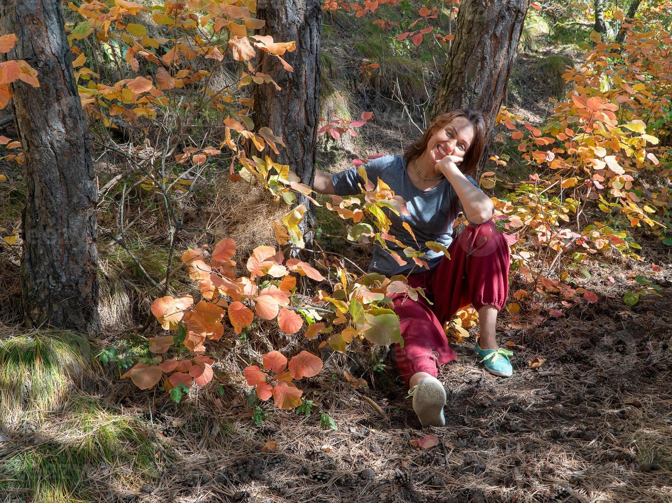 lachende vrouw met rode indische broek zittend in het bos onder de bomen op het pad met herfstgebladerte. portret van een gelukkige vrouw foto