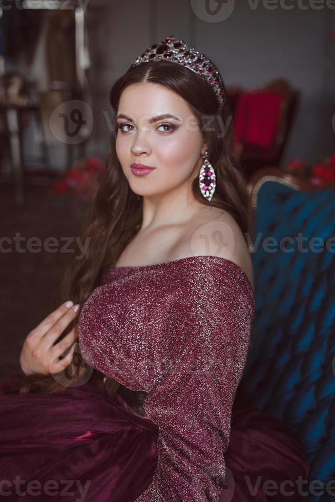 portret van mooie jonge mooie schattige vrouw prinses in lange paarse koningin jurk en kroon, met lang haar en make-up zittend op de blauwe fluwelen bank binnen in minimalistisch interieur foto