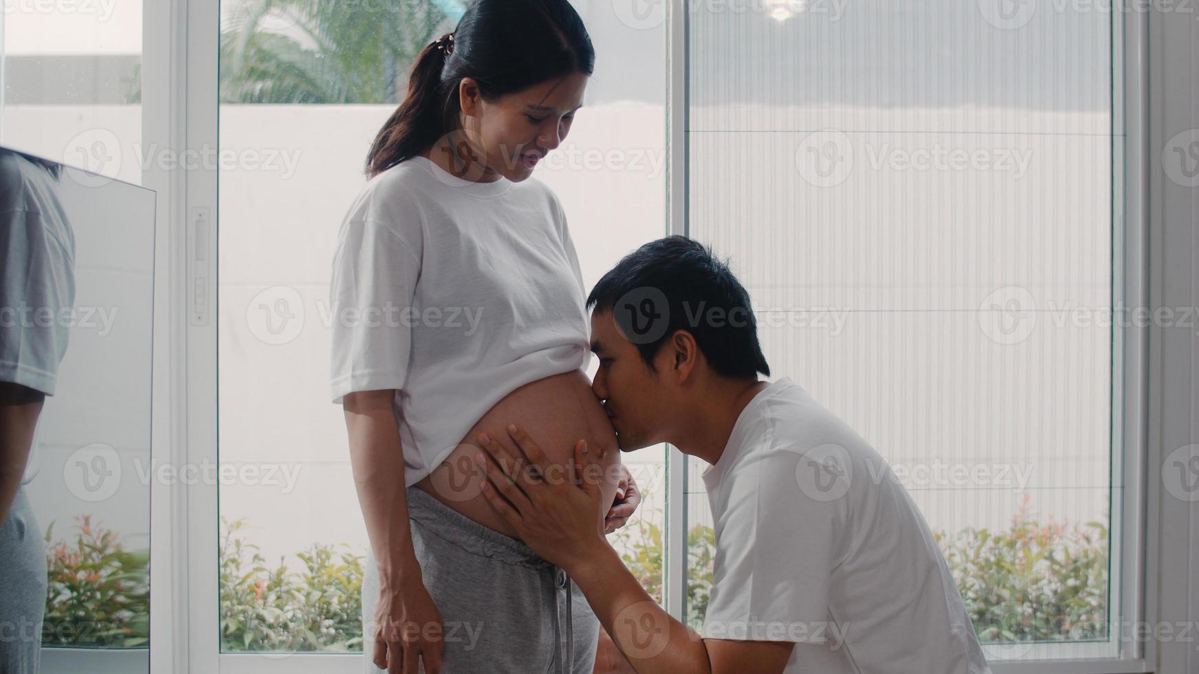 jonge Aziatische zwangere paar man kuste zijn vrouw buik praten met zijn kind. mama en papa voelen zich gelukkig glimlachend vredig terwijl ze voor baby zorgen, zwangerschap bij raam in woonkamer thuis concept. foto