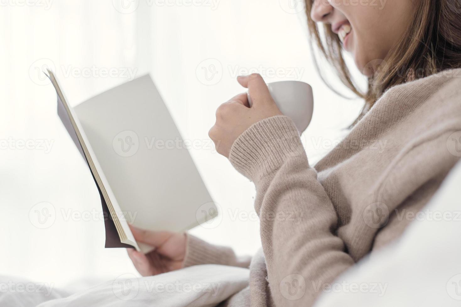 mooie aziatische vrouw die van warme koffie geniet en een boek leest op bed in haar slaapkamer. Azië vrouw draagt een comfortabele trui met een boek en een kopje koffie. levensstijl azië vrouw thuis concept. foto