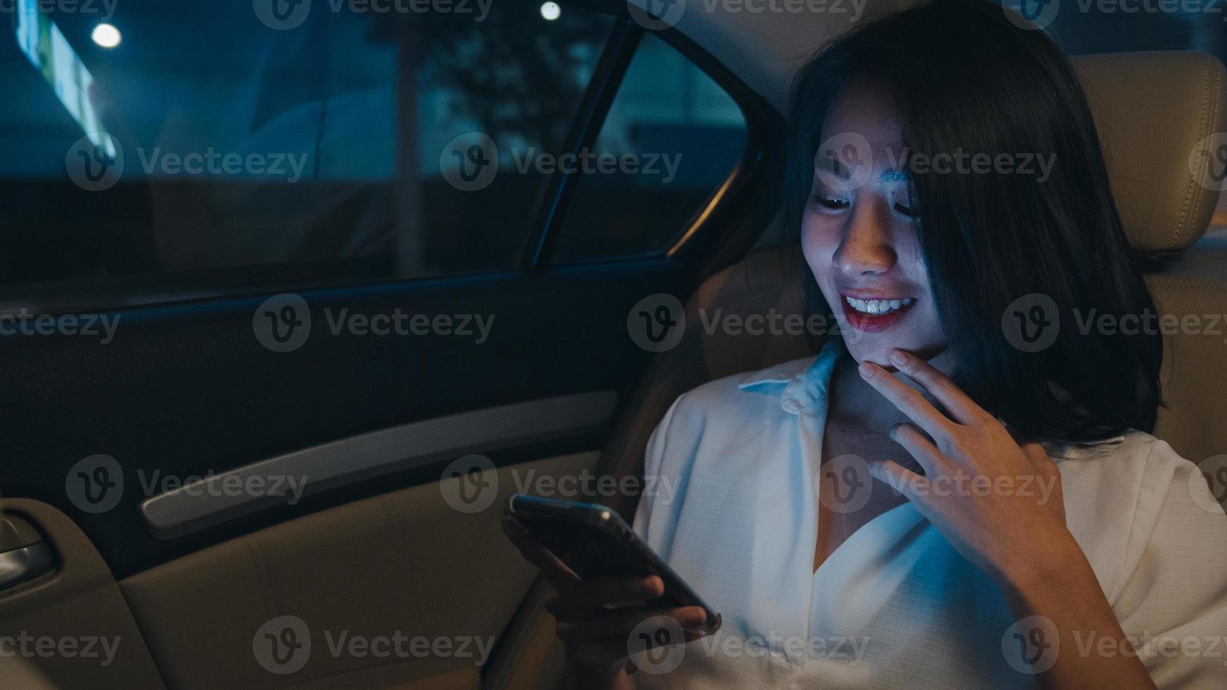 succesvolle jonge azië zakenvrouw in mode kantoorkleding laat werken met behulp van slimme telefoon op de achterbank van de auto in de stedelijke moderne stad in de nacht. mensen beroepsmatige burn-out syndroom concept. foto