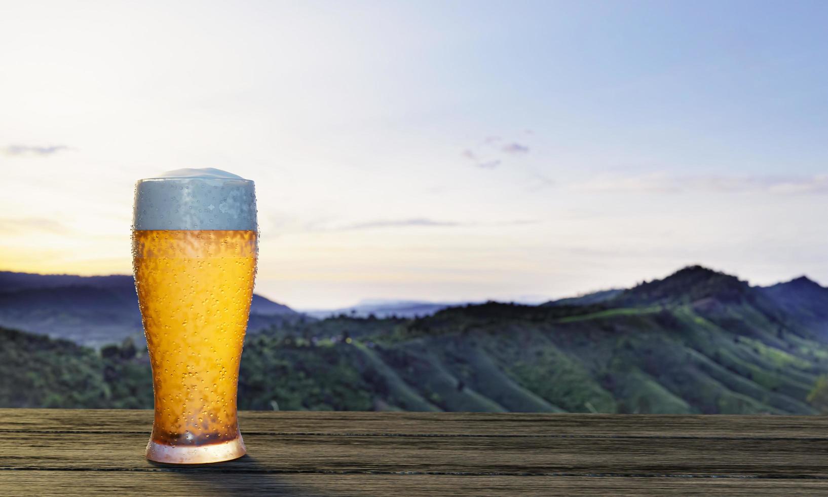 getapt of ambachtelijk bier in helder glas met bierschuim erop op een set houten tafels en stoelen. lounge zitplaatsen op het houten terras met bergachtergrond met godslicht of zonlicht.3D-rendering foto