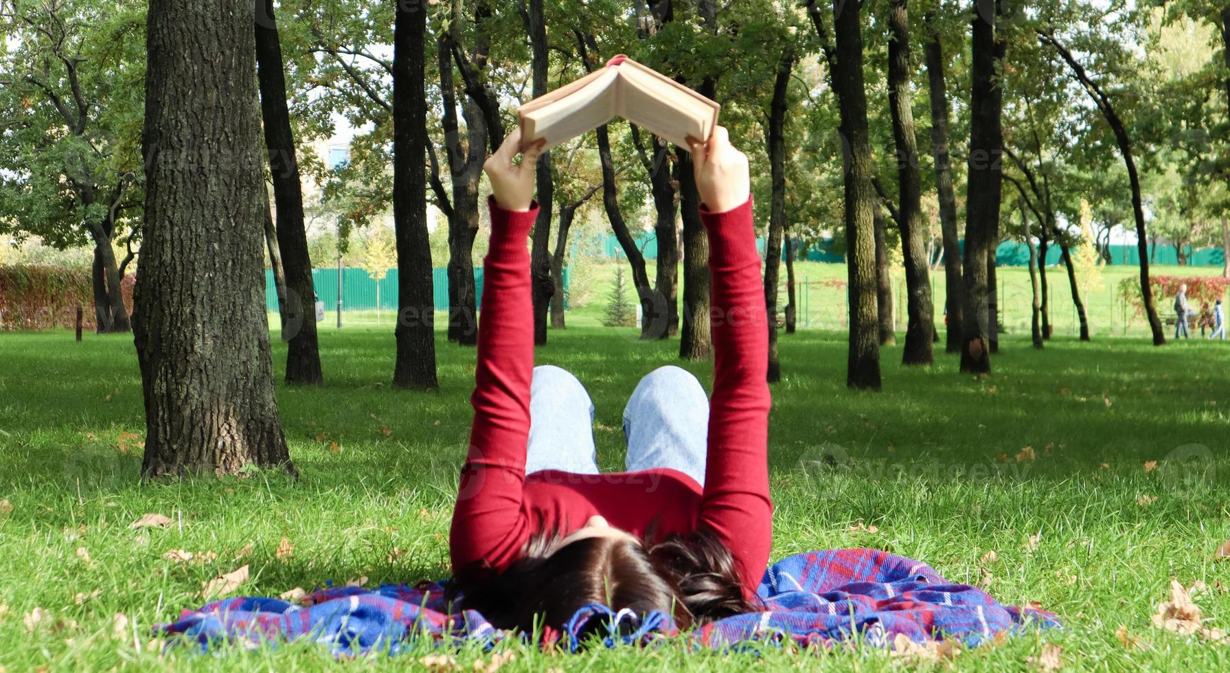 jonge vrouw liegen en lezen van haar favoriete boek op een wollen plaid deken in een stadspark op groen gras op een aangename zonnige dag. het concept van recreatie, onderwijs en studie op afstand. foto