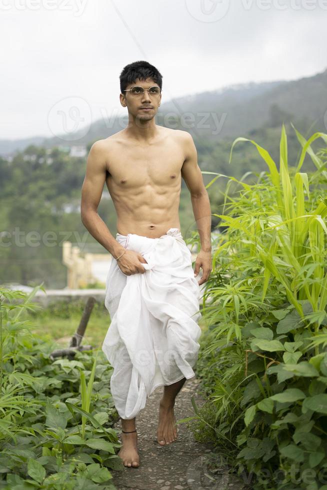jonge indische fitte jongen, lopend op een pad naast gewassen in het veld. een Indiase priester die loopt terwijl hij witte dhoti draagt. Indiase religieuze man. foto