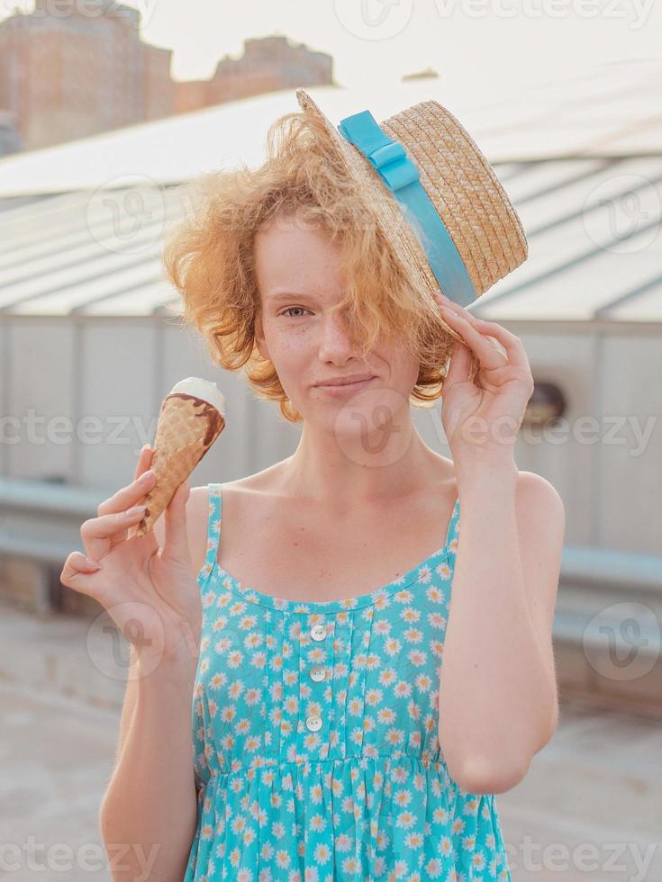 jonge, vrolijke, krullende roodharige vrouw in strohoed, blauwe zomerjurk die ijs eet op het dak van de wolkenkrabber. plezier, lifestyle, stedelijk, modern, dak, stad, zomer, mode, jeugdconcept foto