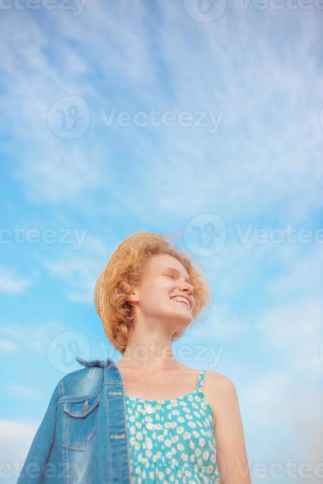 jonge krullende roodharige vrouw in stro hoed, blauwe zomerjurk en jeans jasje staande op blauwe hemelachtergrond. plezier, zomer, mode, schieten, reizen, jeugdconcept. kopieer spase foto
