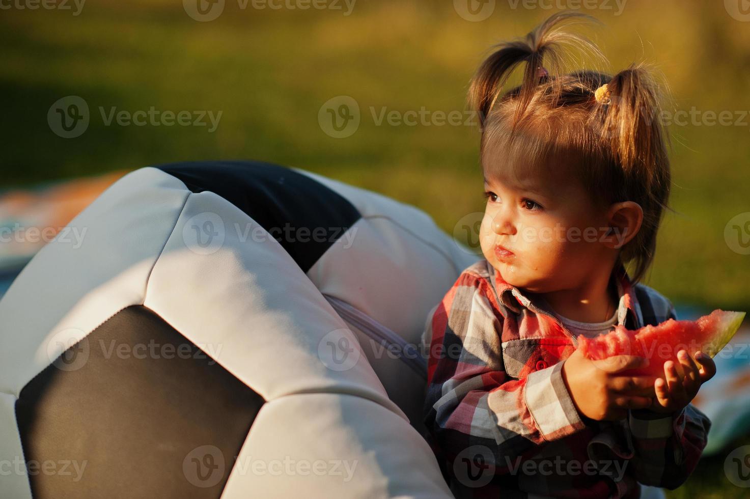 schattig babymeisje in geruit overhemd zit op de poef van de voetbalbal en eet watermeloen. foto