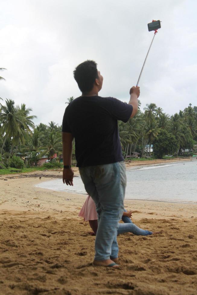 portret van een grote man op het strand die een selfie maakt met een selfiestick. foto